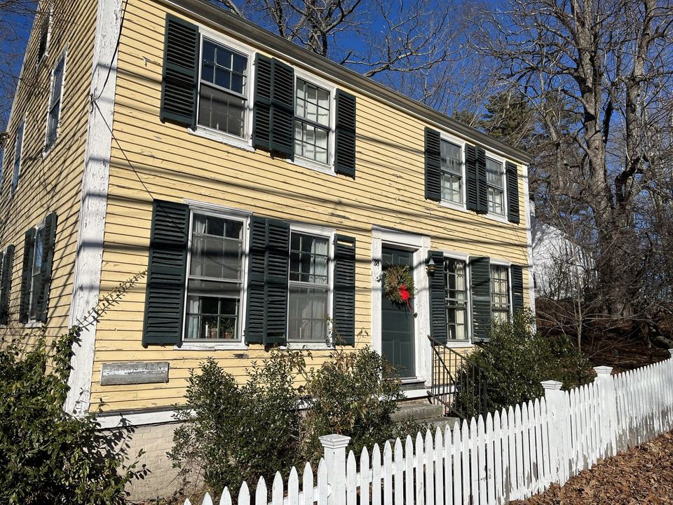 Yellow clapboard house with black shutters behind a white picket fence