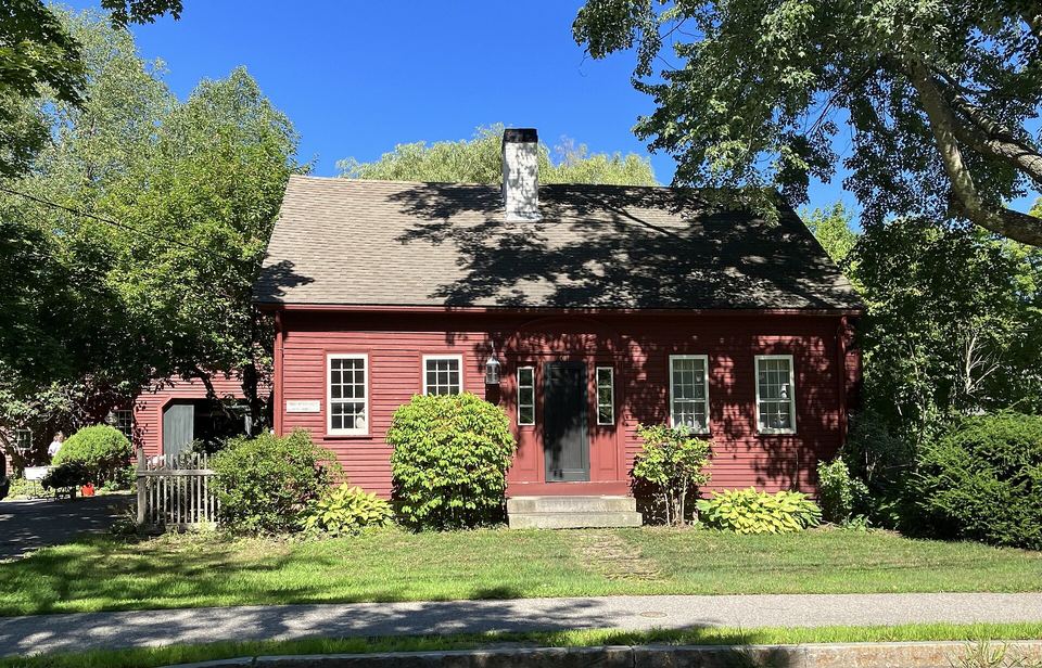 Red clapboard cape-style house with a center chimney, shrubs and a sunny lawn